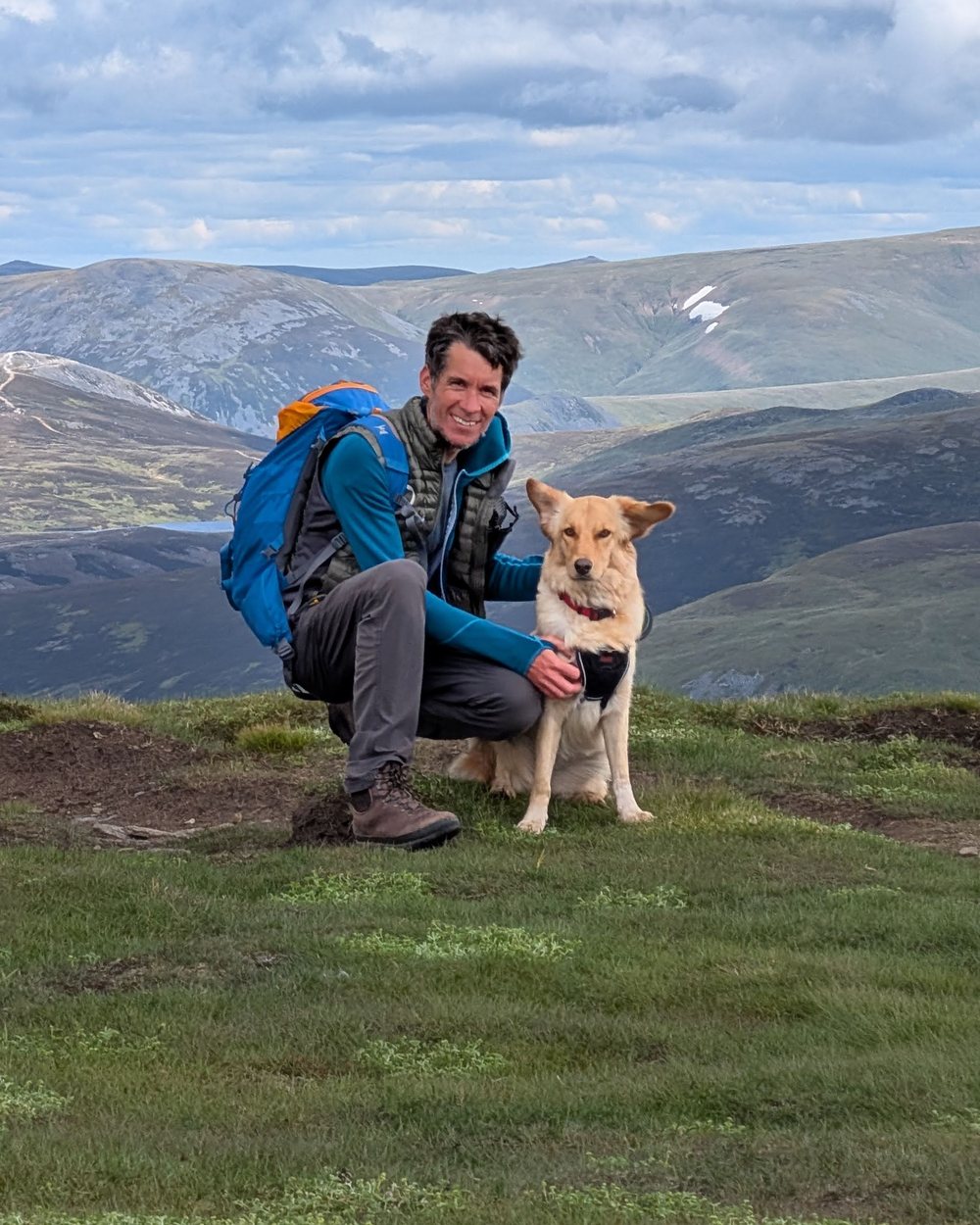 Simon, the maker of Munro Planner, crouching with his dog on a Scottish summit with rolling hills behind